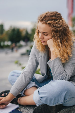 Tired student holding her hand under her head and writing homework in her notebook, sitting outside. Calligraphy, handwriting, creation concept. Business intelligence. Youth lifestyle.の写真素材