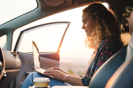 Beautiful curly-haired woman working with laptop sitting on the front seat of a car on a sunny day....