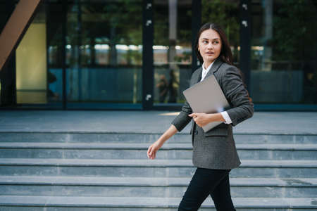 Beautiful business woman in suit holding a laptop in her hands walks down the street. Copy space. Business meeting. Positive person. Business success. Business portrait. City lifestyle.の写真素材