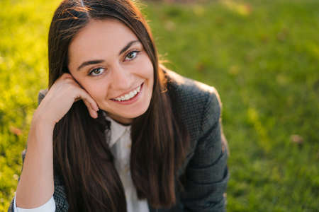 Close-up portrait of a freelancer young woman sitting on lawn in park resting from work in park. Coffee break. Positive person. Smiling and relaxing in summer nature.の写真素材