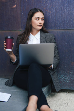 Businesswoman leaning wall outdoors near city building using laptop computer and hold coffee hot cup, working remote online. Beautiful young girl. Education internet technology. Social network.の写真素材