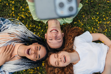 Top view of a group of cheerful bloggers shooting selfie video reading funny comments on website while lying on a grassy lawn. Sunny day and laugh. Emotions and new technologies.の写真素材