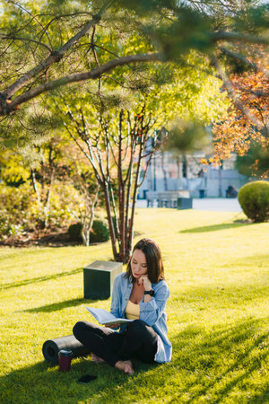 Pretty relaxed woman reading short story, lovely moments, sitting on green grass near yoga mat and...