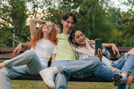 Three happy young teenagers friends having fun at the park, sitting on a bench taking a self portrait with a cellphone. School education. Youth lifestyle.の写真素材