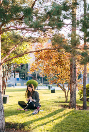 Casual woman sitting on green grass and working part time with laptop and relax in summer camp. Copy space. Drinking coffee to go, freelance using technologyの写真素材