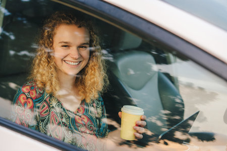 View from the car window of a female student studying online on a laptop holding a coffee while they...