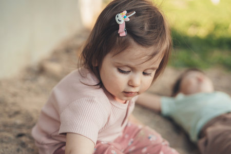 Close-up portrait of a baby girl sitting in the sand in the garden with her brother, exploring. Summer activities for children. Sunny summer dayの写真素材