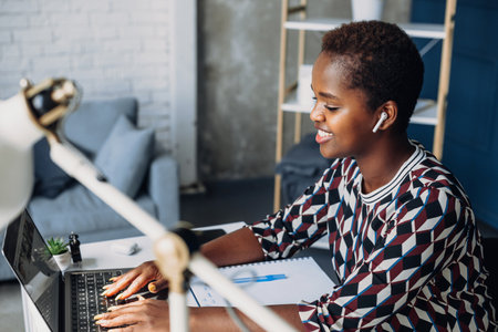 Smiling young African American businesswoman working on a laptop at her desk in office. Remote job, technology and people conceptの写真素材