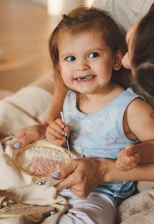 Woman with her little daughter sitting on the floor embroidering with a cross-stitch thread. Parent,...