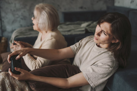 Excited senior mother and mature daughter sitting on the floor, relaxing playing video games at home together, in living room. Happy family together. People having fun on bright living room.の写真素材