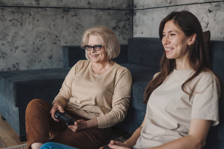 Happy adult daughter and senior mother relaxing on the couch playing video games at home in the...