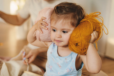 Funny portrait of a baby girl holding two large balls of thread to her ears, the texture of the thread being very soft and pleasant to the touch. Fabric baby design. Child care. Creative design.の写真素材