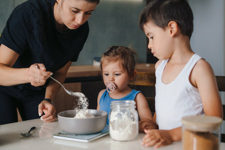 young mother with her two kids weighing out flour into bowl in kitchen environment, baking cakes for the party. Weighing indigrients on electronic kitchen scalesの写真素材