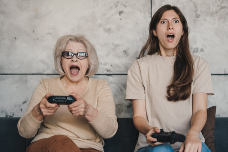 Caucasian senior woman with her young adult daughter having fun enjoying play video game funny video using joysticks at home sitting on sofa. Technology and lifestyle concept.の写真素材
