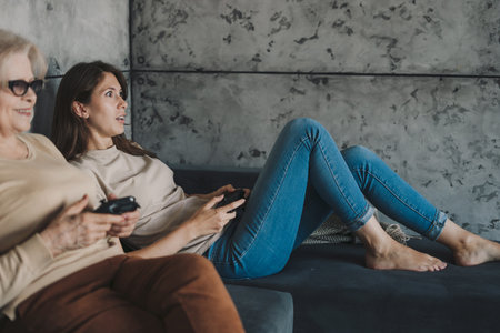 Portrait of overjoyed different female generations family playing video games at home, sitting on sofa in their free time. Modern for video game design. Happy family.の写真素材
