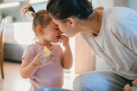 Caring Caucasian mother kissing her little girl on the nose while the little girl eats a cookie. Cute little girl with a cookie in her mouth. Childrens emotionsの写真素材