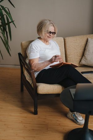Positive senior woman wearing eyeglasses working remotely while sitting on sofa with laptop computer...