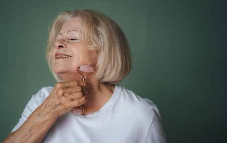 Beauty face portrait of pretty senior woman massaging her face with a jade roller, closing her eyes, isolated over green wall. Natural anti wrinkle facial technique and wellness routine. Senior faceの写真素材