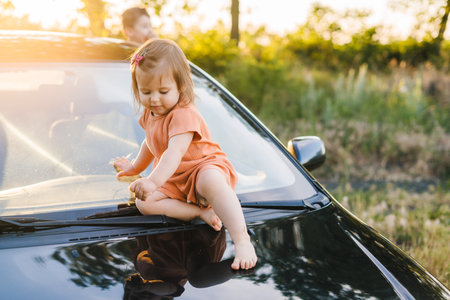 Kid little child baby girl sitting alone without supervision on the hood of the car. Smiling happy child. Happy family, childhood. Summer vacation fun. Travel trip.の写真素材
