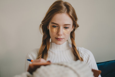 Beautiful young woman with under eye patches holding pen with notebook and sitting on sofa at home office. Education concept. Relaxing time.の写真素材
