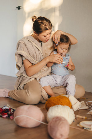 Smiling young Caucasian mother sitting on the floor, knitting with needles with her cute little baby girl daughter. Improve motor skills with small girl child at home. People lifestyle. Fun family.の写真素材