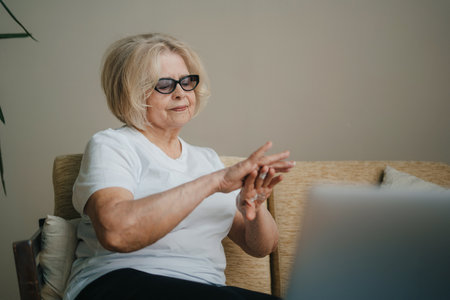 Elderly woman applying cosmetic cream onto her hands while taking a break from working on the computer. Beauty treatment.の写真素材