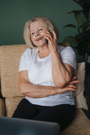 Senior woman with eye-patches talking on phone and using laptop in living room. Family home leisure. Internet communication, wireless connection technology.の写真素材