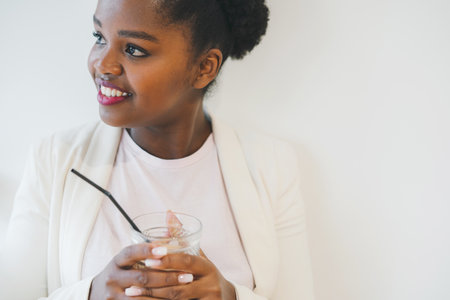 Young black female sitting in street cafe with glass of coffee beverage while waiting friends. At...