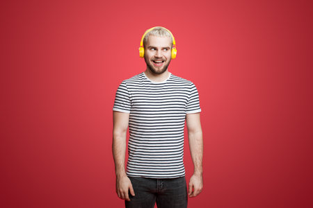 Portrait of amused and happy young guy listening music in headphones, smiling pleased, standing over...