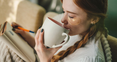 Pleasant young caucasian woman sitting on the sofa and drinking a mug of coffee wearing under-eye...