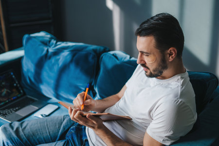 Portrait of concentrated man using laptop for remote work sitting on blue sofa, holding pencil, noting down in notepad. Occupation and technology.の写真素材