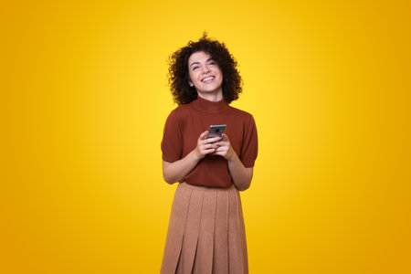 Portrait of young curly-haired woman using smartphone for messaging with friends standing over yellow studio background.の写真素材