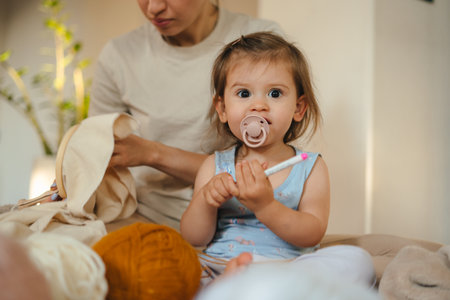 Pretty baby girl sitting near her mother doing housework during quarantine, embroidery with colored...
