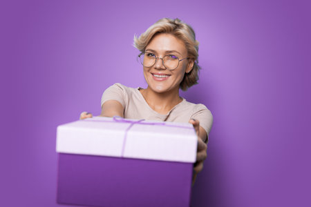 Beautiful young woman giving gift box at camera on purple studio background. Lovely millennial lady offering present with smile, celebrating special occasionの写真素材
