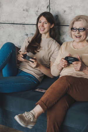 Adult daughter with her senior mother playing video games with console controller sitting on sofa at home. Activity relationship. Family home leisure. Happy family.の写真素材