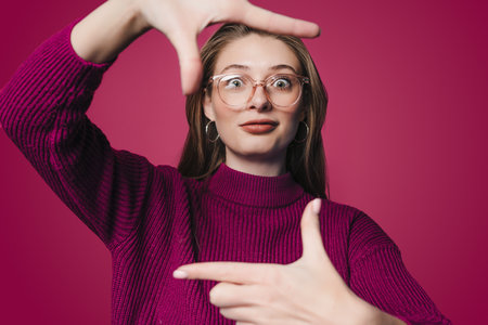 Young beautiful woman smiling, looking at camera and making frame with hands and fingers standing over crimson background. Creativity and photography concept.の写真素材
