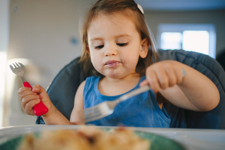 Front view of a hungry baby girl sitting in her high chair at home, eating breakfast by fork feeding herself.の写真素材