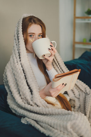 Thoughtful and cozy woman drinking morning coffee sitting on sofa wrapped in a blanket and reading a...