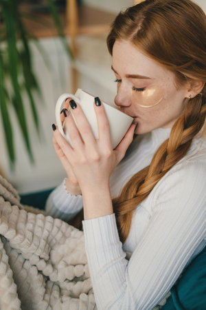 Portrait of attractive red-haired charming woman wrapped in soft blanket sitting on sofa drinking...