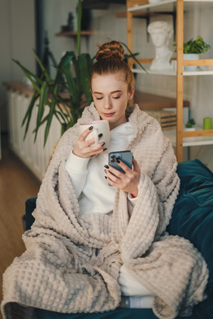 Happy beautiful young woman with a smartphone and cup in hands wrapped in a plaid on the couch while resting at home. Winter season.の写真素材