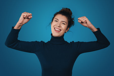 Excited young woman with raised hands and fists opening mouth and screaming while celebrating success isolated over blue background. Girl get happy, enjoy life.の写真素材