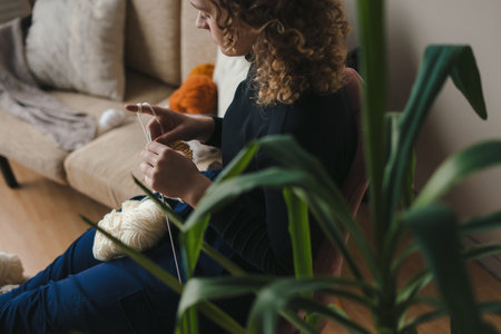 Over shoulder portrait woman knitting clothes relaxing in big chair. Cozy interior with young female. Cozy home concept.の写真素材