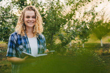Curly-haired woman farmer writing notes on clipboard notepad in organic farm orchard.の写真素材