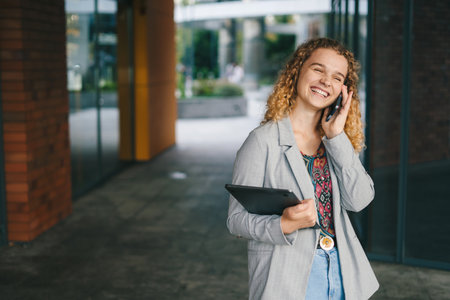 Positive young female wearing jacket standing outside with laptop and making phone call while looking away and laughing. Student standing call outdoors. Holding smartphone.の写真素材