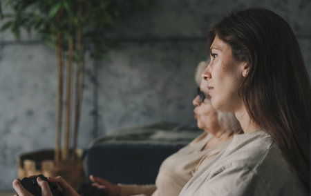 Side view of a woman with her mother playing games on TV, sitting on cozy sofa. Healthy activity. Happy cheerful family. Happy lifestyle. Family home leisure.の写真素材