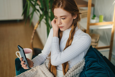 Red-haired woman texting message on smartphone sitting on sofa at home. Rest, relax on vacation in bedroom. Reading news in social media, browsing, surfing in internet.の写真素材
