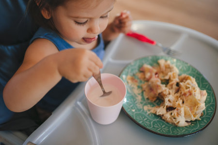 Caucasian baby girl eating lunch from plate sitting high chair, self feeding. Healthy nutrition of solid food for infant. Learning concept.の写真素材