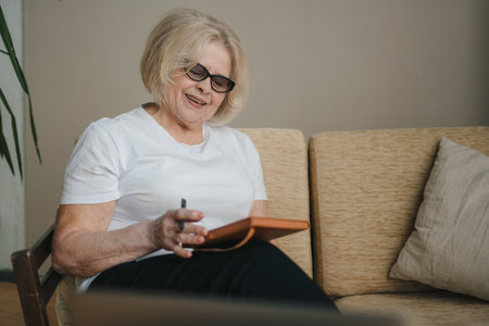 Photo portrait of elder woman using laptop computer watching webinar sitting on sofa writing in notebook. Happy stylish mature woman remote working from home. People lifestyle portrait. People using laptop.の写真素材