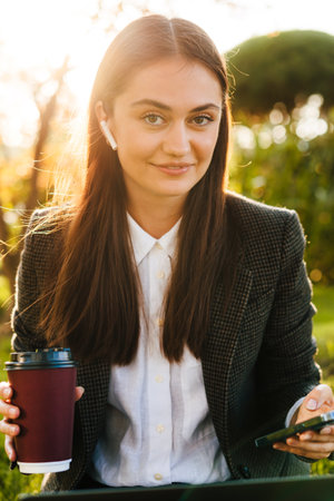 Portrait of a young adult caucasian business woman using mobile phone in city park, drinking coffee cup while looking at camera. Sitting woman outdoors. People using laptop.の写真素材