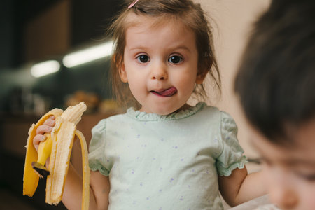 Happy portrait of a cute little baby girl holding peeled banana for eating, sticking out tongue, looking at camera. Healthy food. Happy family. Sweet food.の写真素材